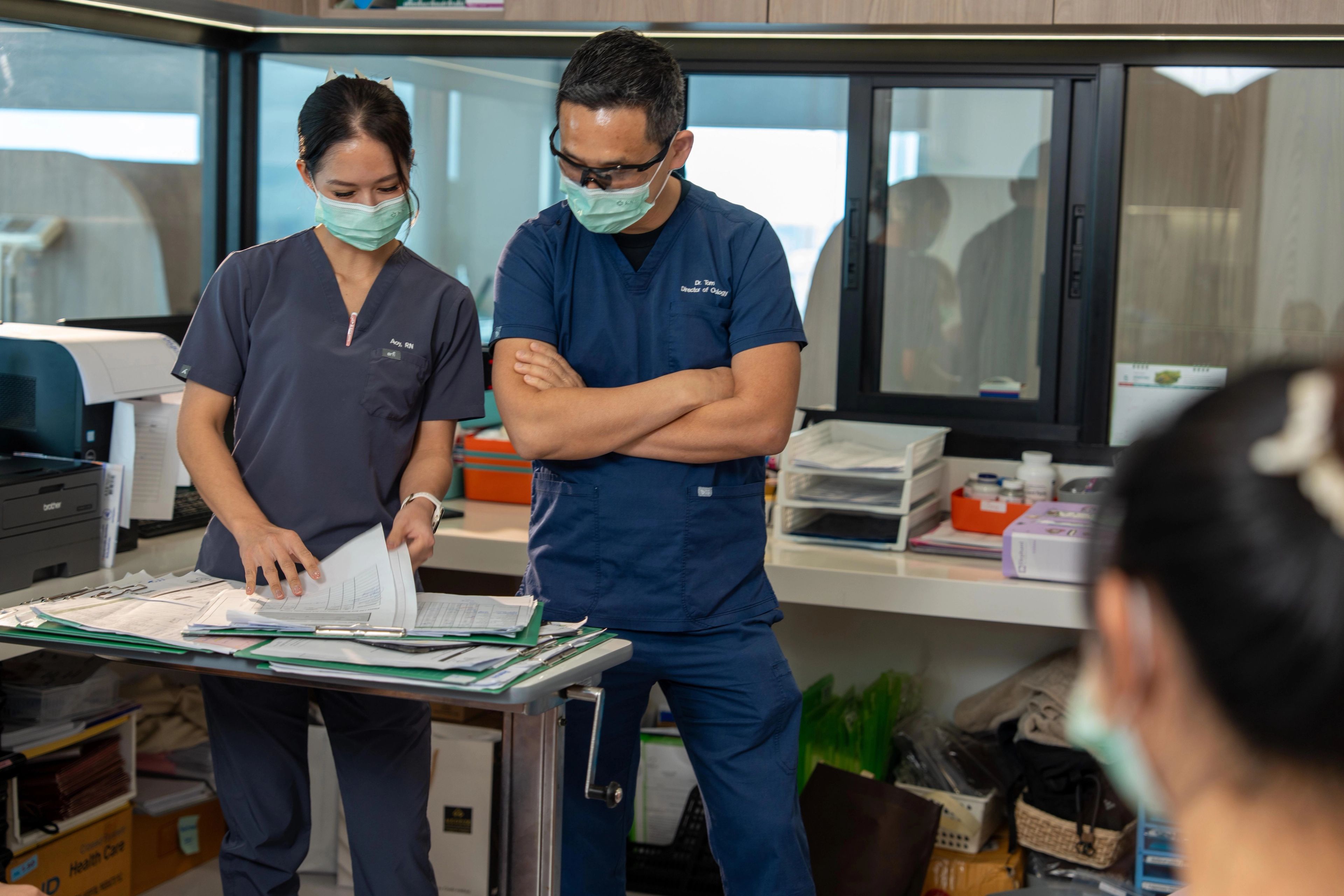 Nurse and doctor in scrubs and face masks having a conversation while looking at notes on a desk in an office, there is a third medical professional in the room.