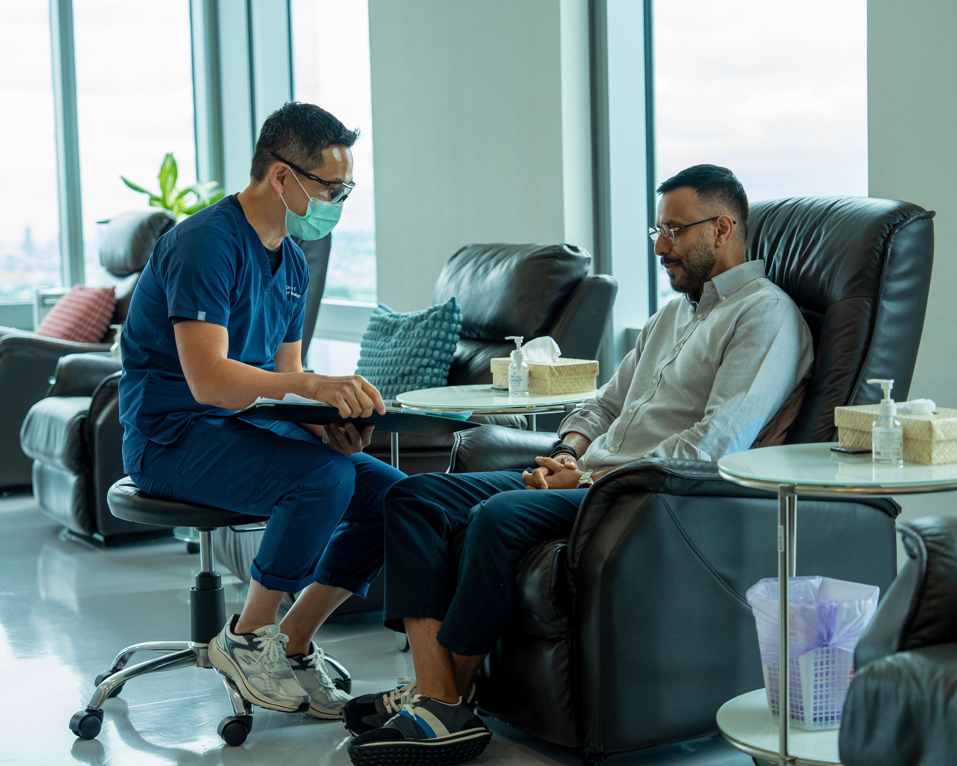 Doctor in navy scrubs and a surgical mask sits in front of a patient in a black leather chair having a conversation.