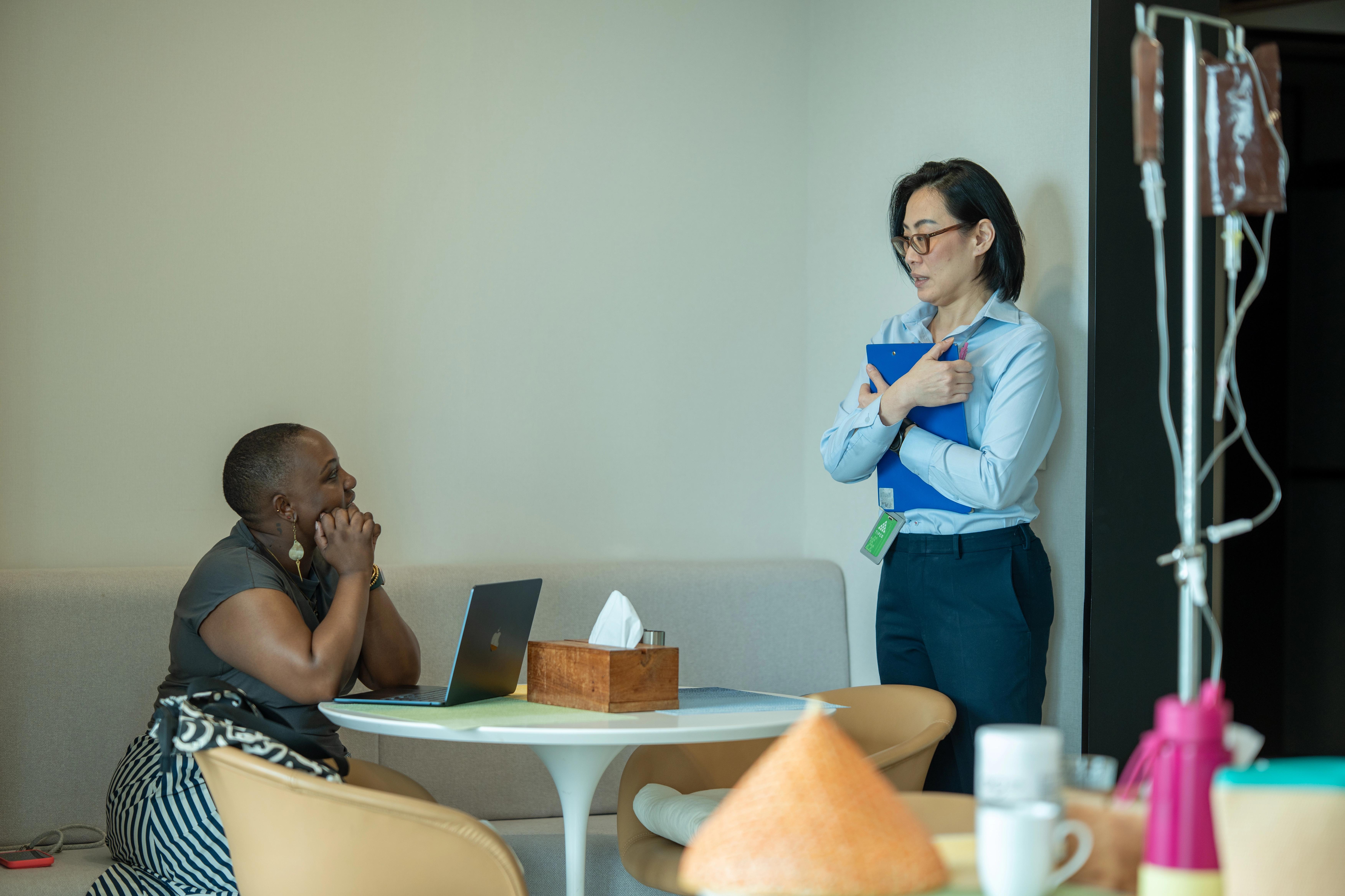 A patient seated at a table in front of a laptop, having a conversation with a woman standing holding a folder. You can see a saline drip out of focus.