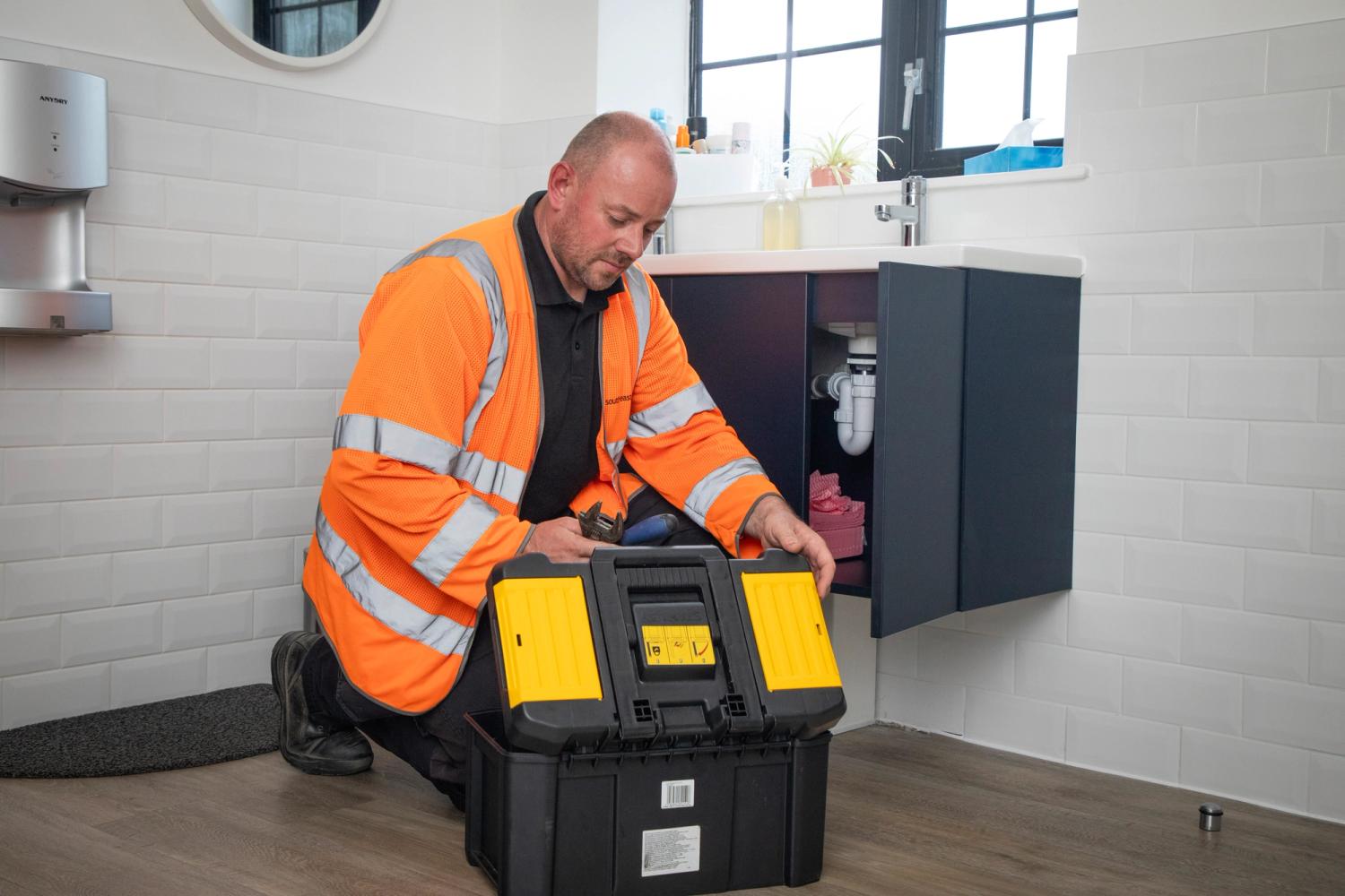 Man in a hi-vis looking under a sink with a black and yellow toolbox at his side.