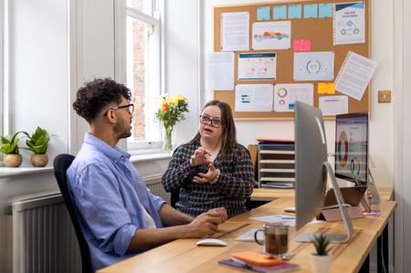 Two people chat around a computer in an office setting