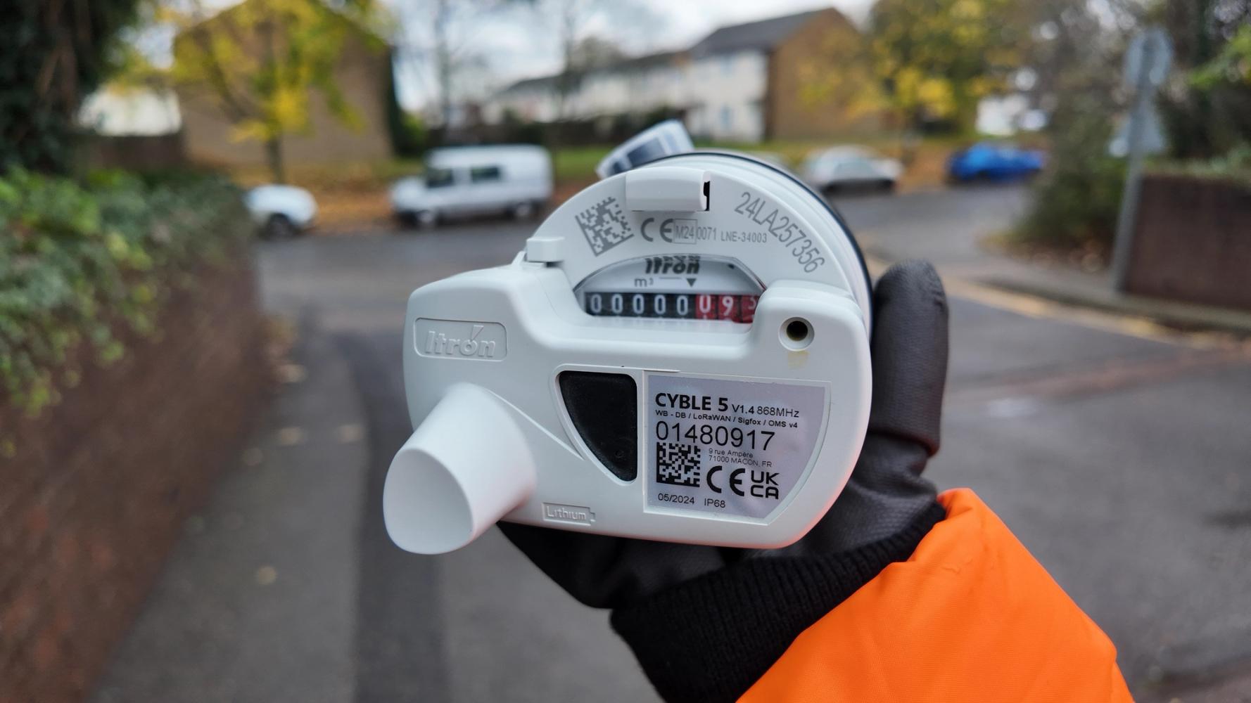 A close-up of a technician's gloved hand holding a white meter with a visible reading of 0000009. The background shows a blurred residential street with parked cars and houses.