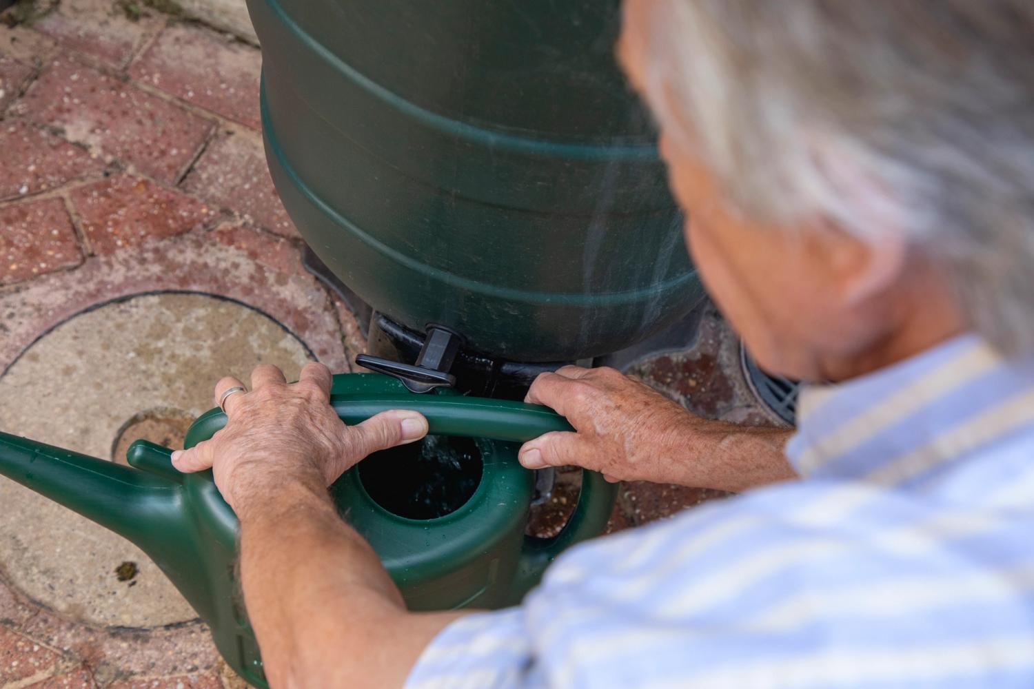 A person using a green watering can to collect rainwater from a water butt.
