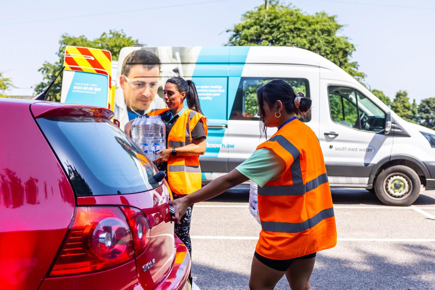 Photo of 2 SEW employees wearing orange high vis at a bottle water station. Loading bottles of water into the boot of a red car.