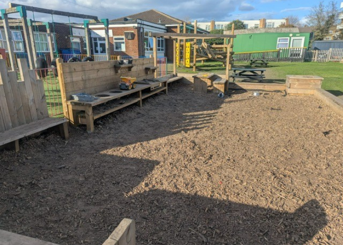 Image of the current sensory garden at St Joseph's school which shows an outdoor play area covered in wood chips, featuring a wooden mud kitchen structure with a toy yellow dump truck on the counter.
