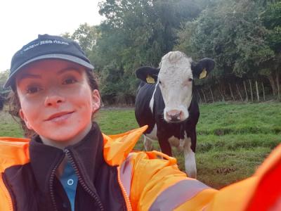 A South East Water Catchment Scientist, Tauba, wo is wearing a South East Water hat, hi-vis jacket, standing in front of a cow in a field.