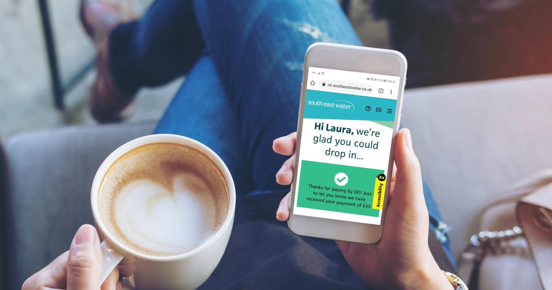 Photo taken from above of a person sat on a sofa holding a cup of coffee in their left hand and a mobile phone in their right hand