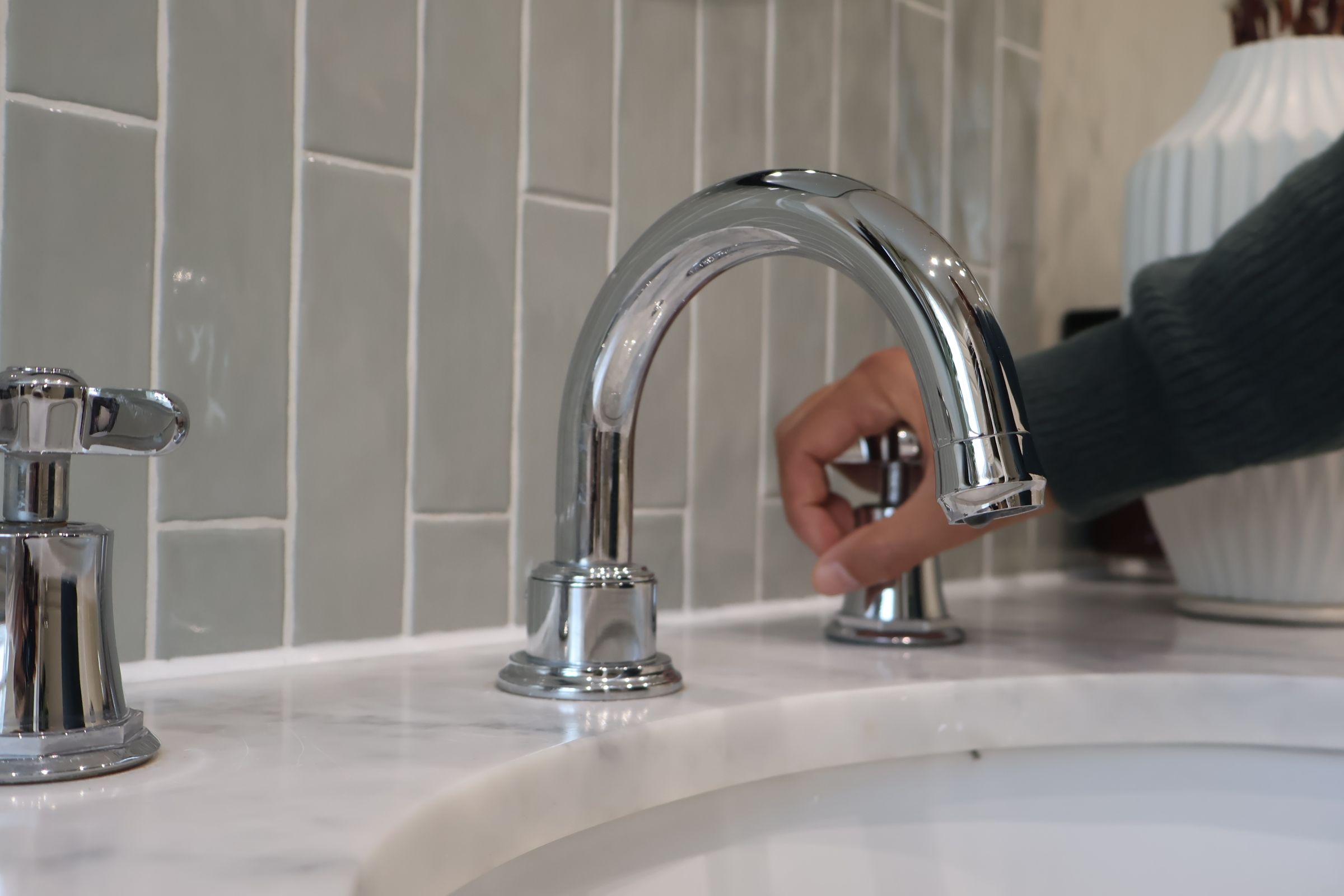 An image of a sink within a stone worktop. A person has their hand on the cold tap. There is no running water. 