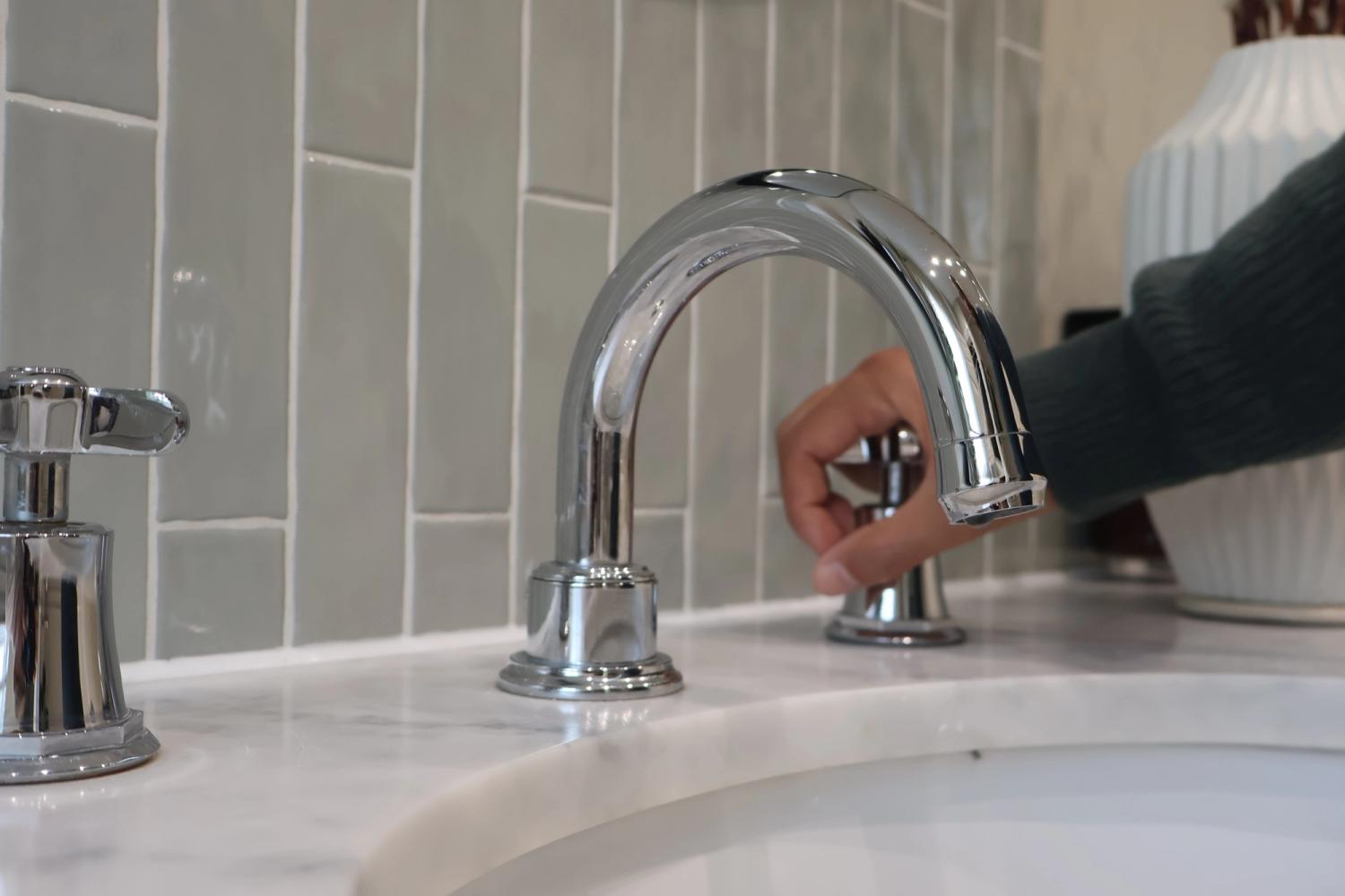An image of a sink within a stone worktop. A person has their hand on the cold tap. There is no running water. 