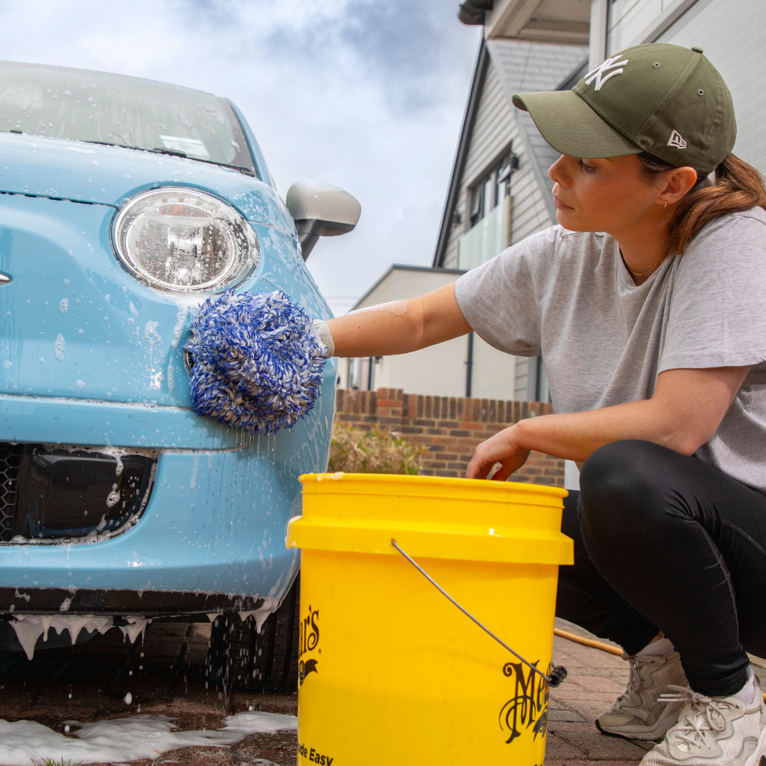 A women using a yellow bucket filled with water to wash her light blue car with a sponge.