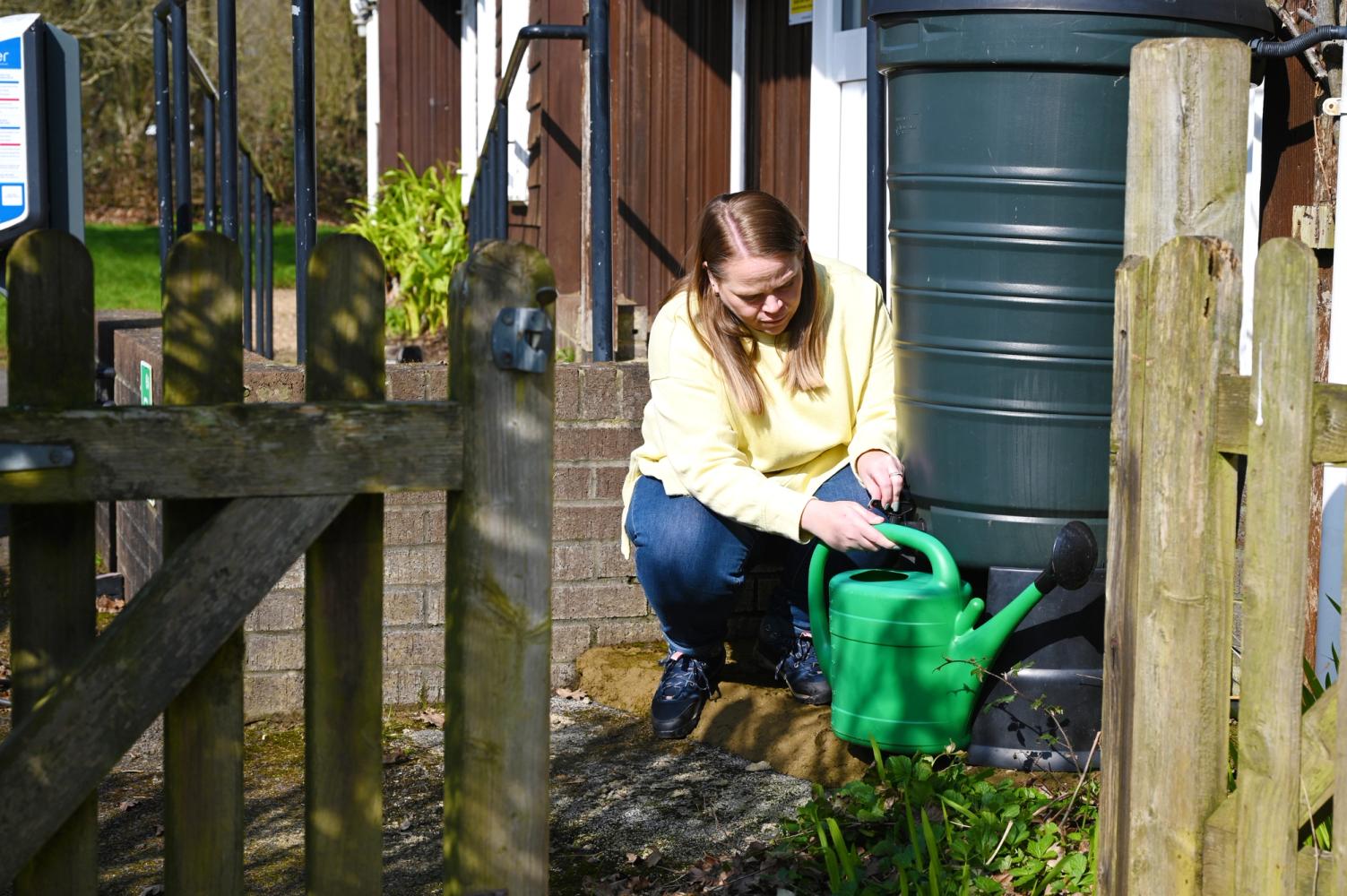 A women bending down filling up a light green watering can from a dark green water butt.