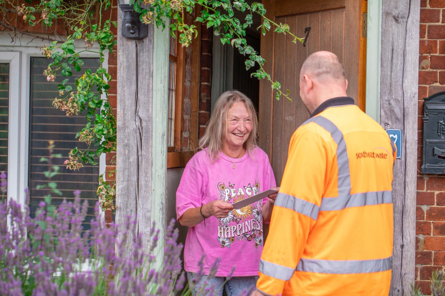 A South East Water employee wearing an orange his-vis jacket is stood chatting to a customer at her front door. He is handing her a leaflet.