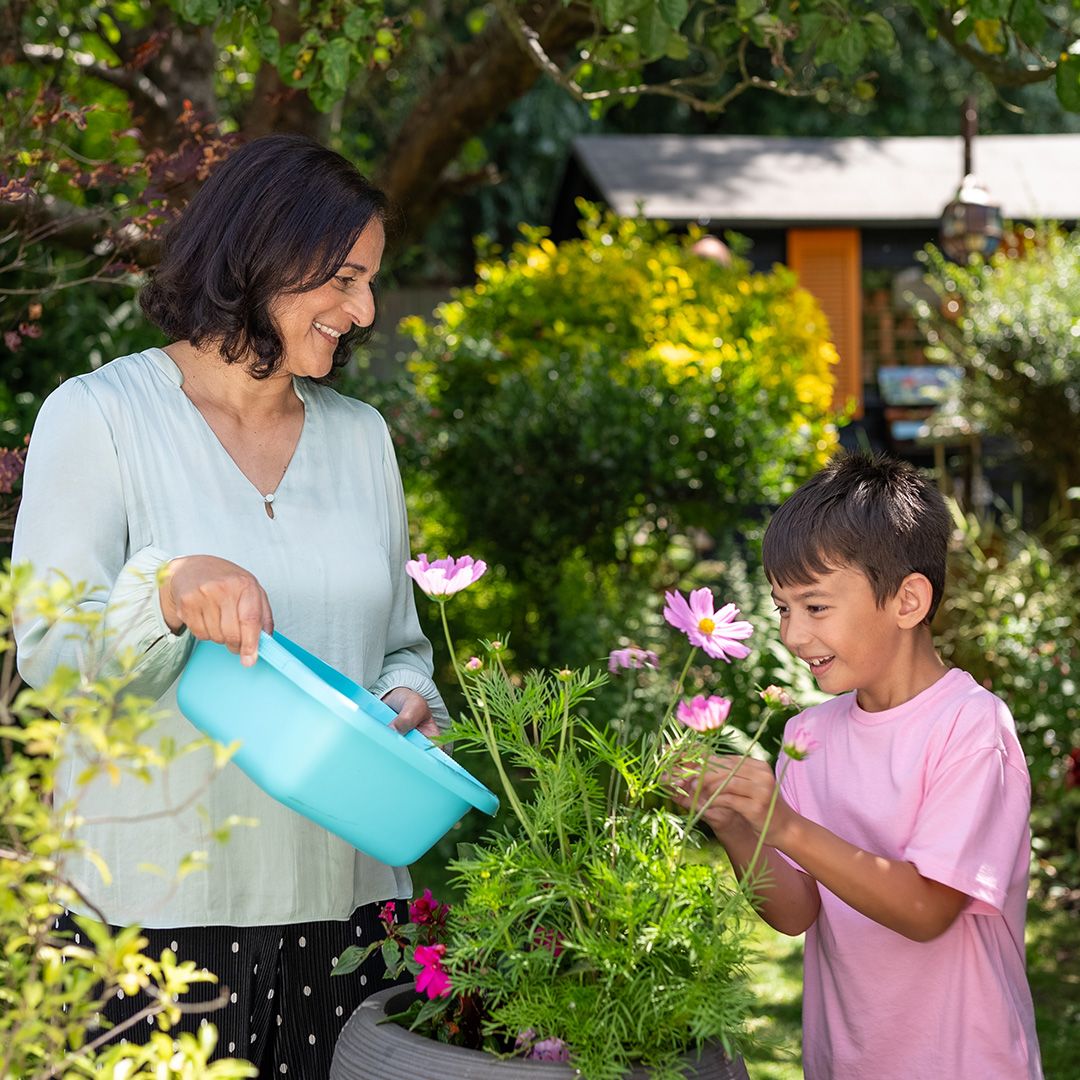 Photo of a grandmother reusing sink grey water from a washing up bowl to water her plants, with her grandson. 