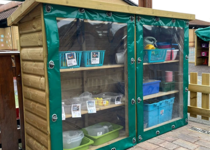 An image of a wooden, three-tier outdoor school storage unit with a sloped roof. The front is protected by two clear PVC covers with green edges, which are fastened shut. Inside the shelves are various colourful plastic storage tubs and children's play items.