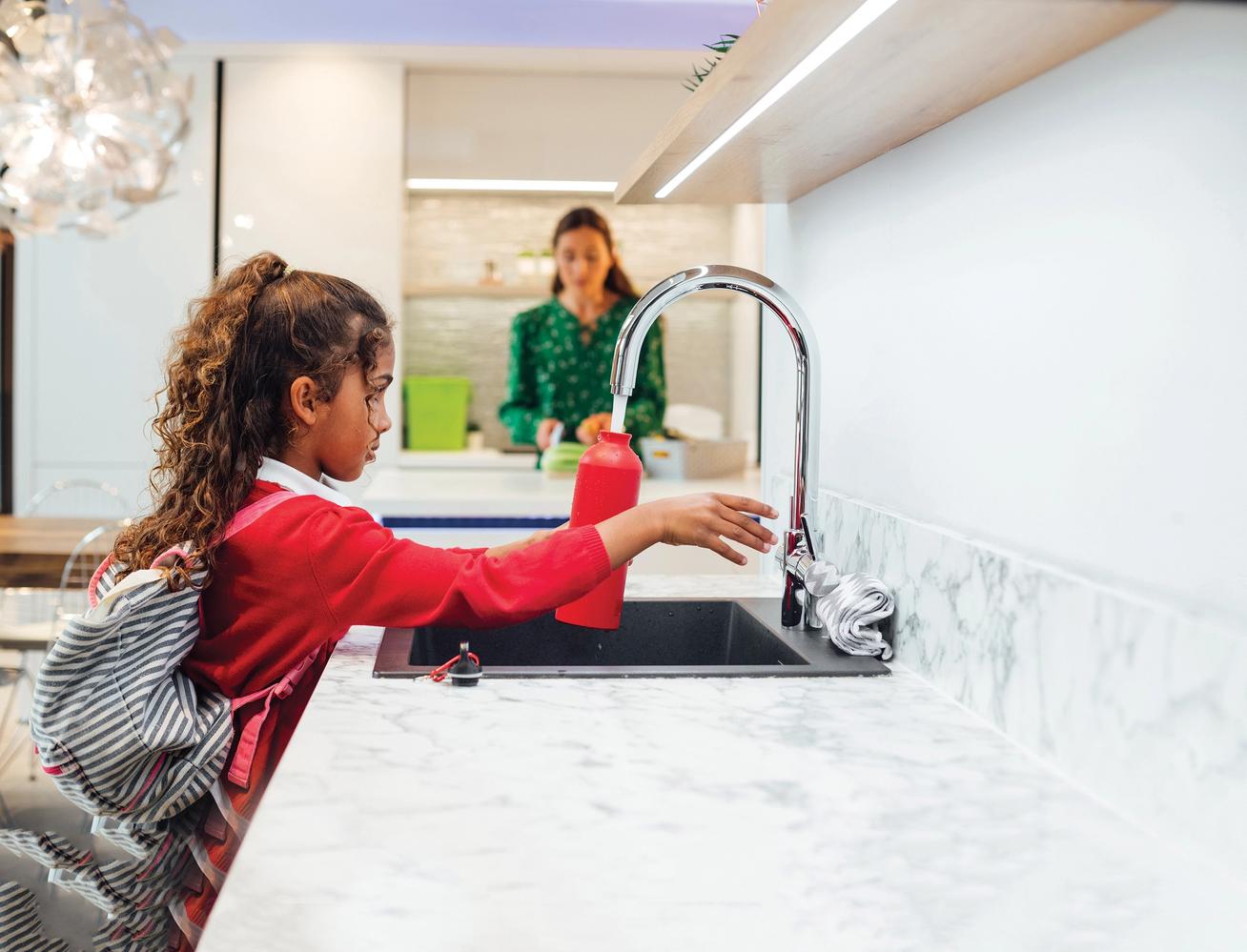 A young girl wearing her school uniform filling up her water bottle at the kitchen sink