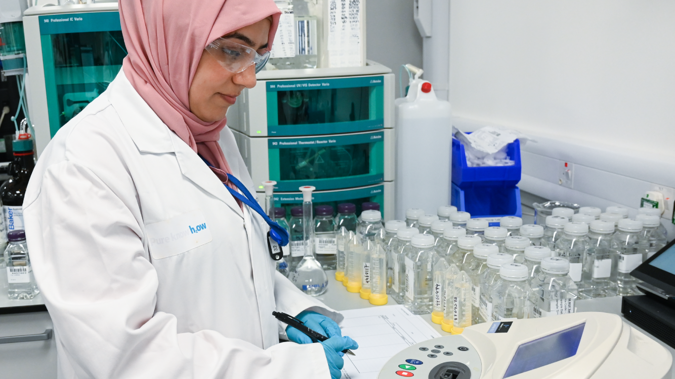 Photo of a South East Water employee wearing a lab coat, writing notes in a laboratory. Bottles of water to be tested and equipment can be seen in the background. 