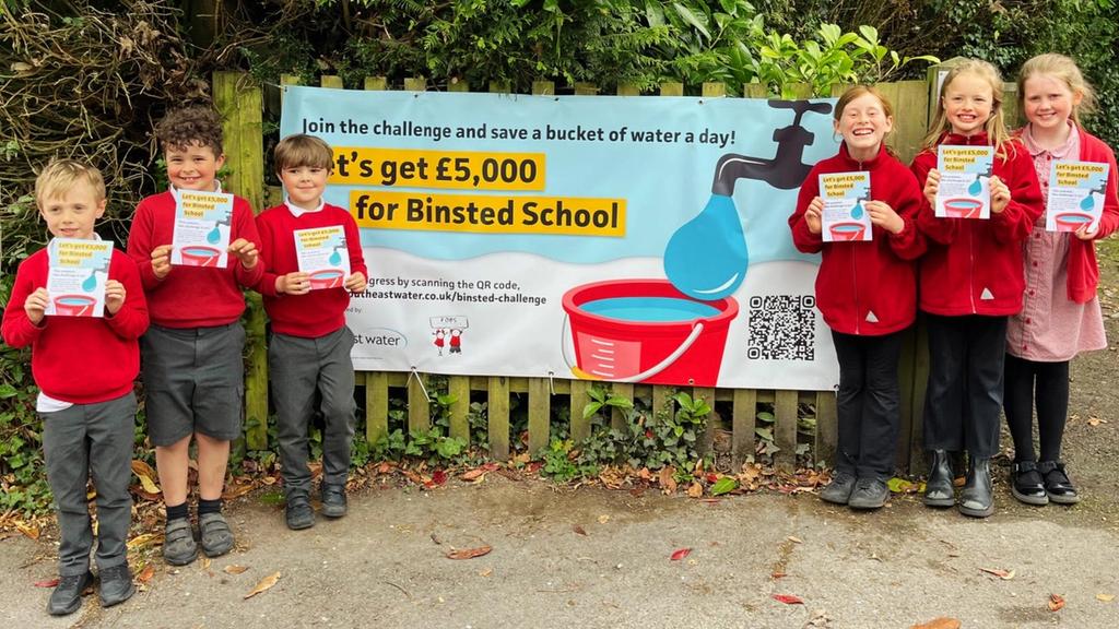 Pupils of Binsted School holding a challenge leaflet and stood beside the challenge banner attached to the school fence.