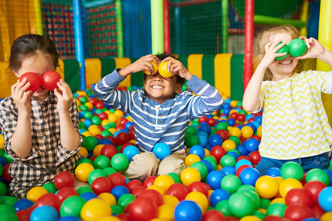 Three children in a ball pit, each covering their eyes with plastic balls from the ball pit. 