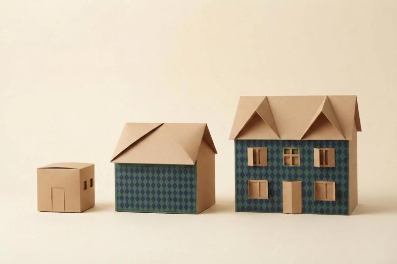 Paper-craft illustration of three folded kraft paper homes in rising sizes, each with its own small stack of paper cartons beside the door.