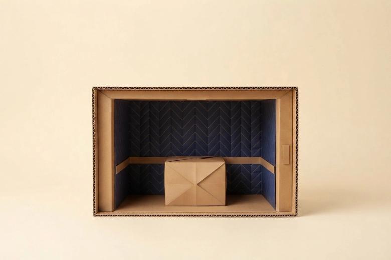 Paper-craft illustration of a folded-paper elevator car with quilted paper pads hung inside, doors held open, and a small kraft paper box waiting in the corridor