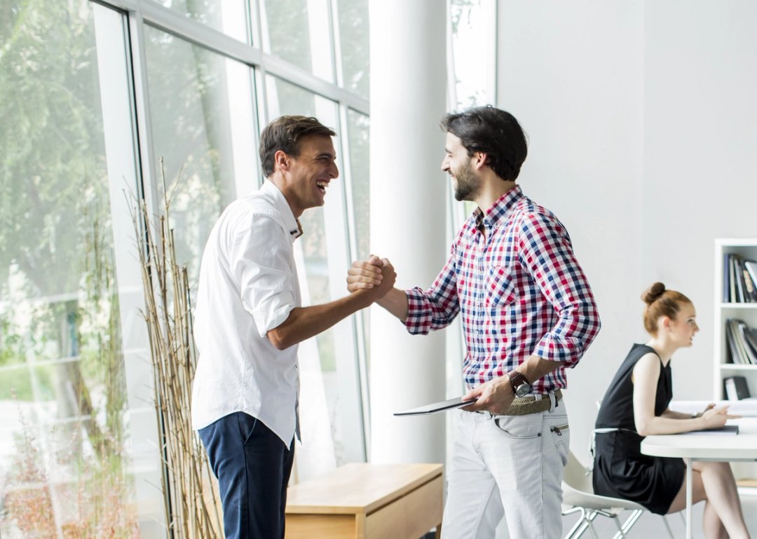 Zwei Männer reichen sich lachend die Hand in einem hellen Büro