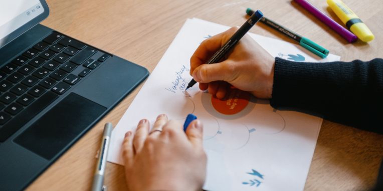 Hand zeichnet mit Stift ein Diagramm auf Papier neben einem Tablet mit Tastatur