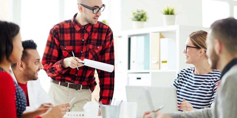 Teambesprechung am Tisch, ein Mann steht mit Unterlagen und spricht zur Gruppe