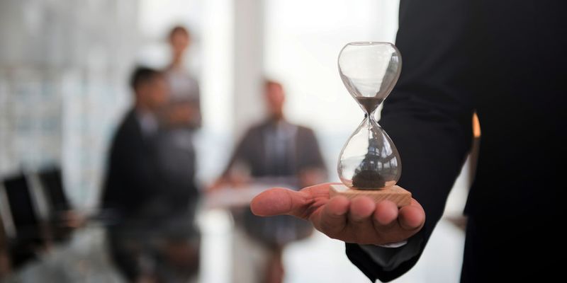 Hand hält eine Sanduhr, im Hintergrund unscharf ein Besprechungsraum