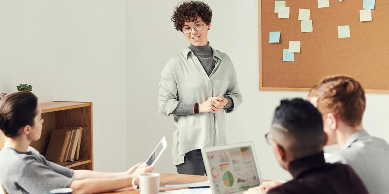 Frau präsentiert vor einer Pinnwand, Team am Tisch mit Laptop und Tablet hört zu