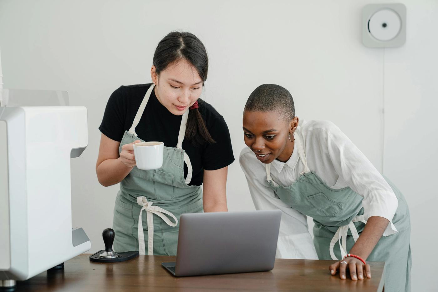 Zwei Mitarbeiterinnen in Schürzen schauen gemeinsam auf einen Laptop in einem Café
