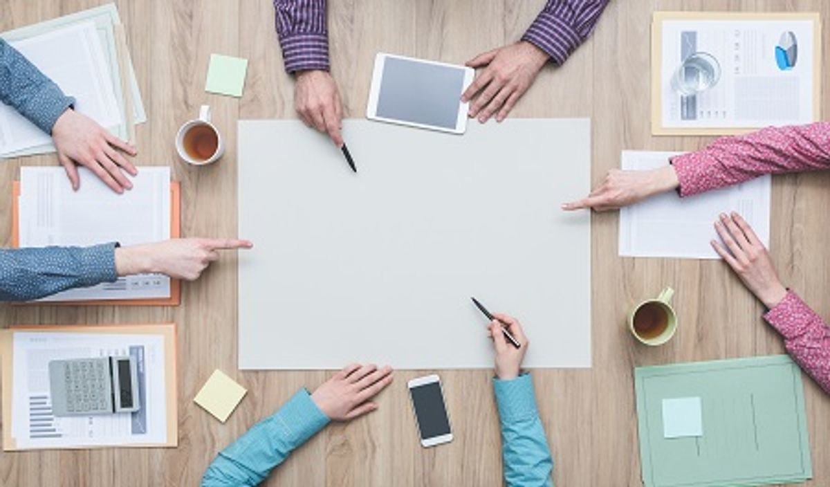 A group of hands pointing at a blank sheet on a table, surrounded by documents, a tablet, and cups of coffee.