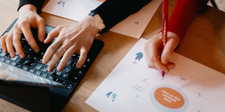 Hände tippen auf einer Tastatur, daneben skizziert eine Person eine Mindmap auf Papier