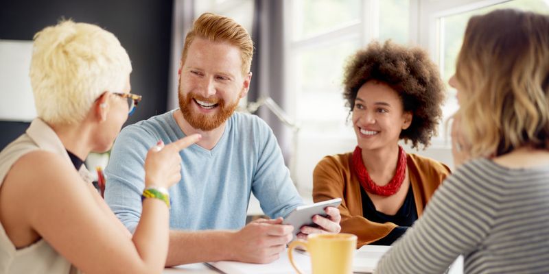 Vier Kolleginnen und Kollegen unterhalten sich lachend am Tisch mit Tablet