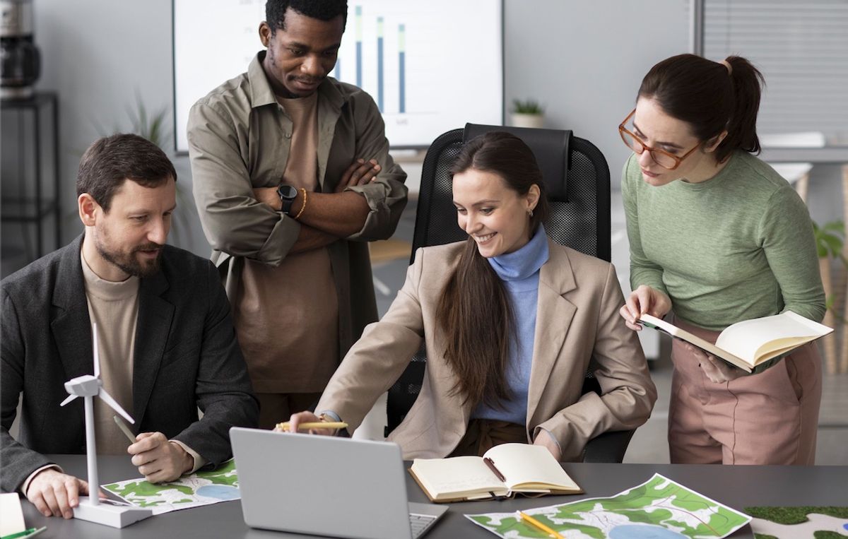 A group of four professionals collaborates around a laptop, discussing plans with maps and notebooks on a modern office table.