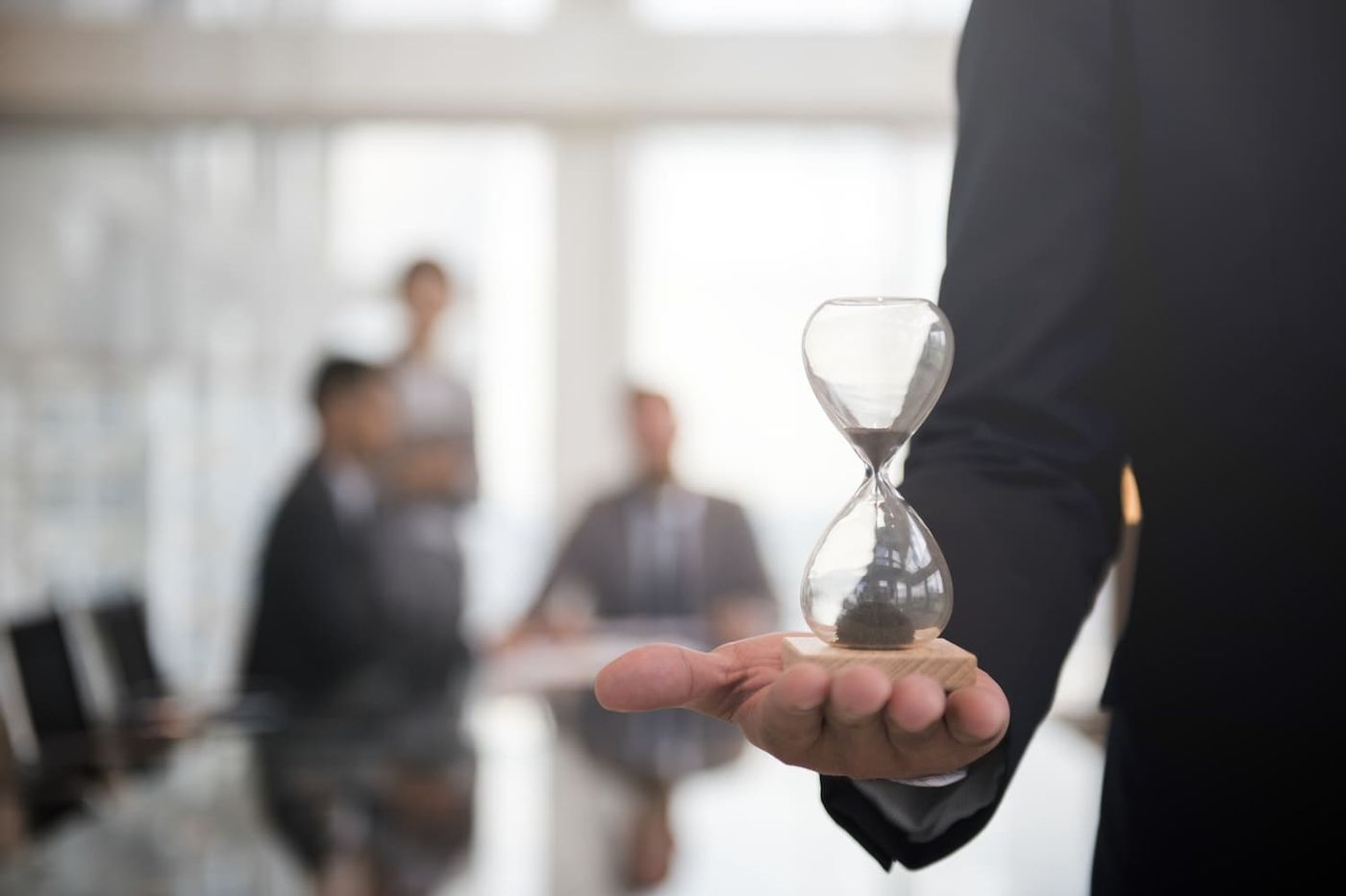 Person hält eine Sanduhr auf der Hand, im Hintergrund ein Team am Besprechungstisch