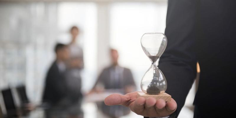Person hält eine Sanduhr auf der Hand, im Hintergrund ein Team am Besprechungstisch