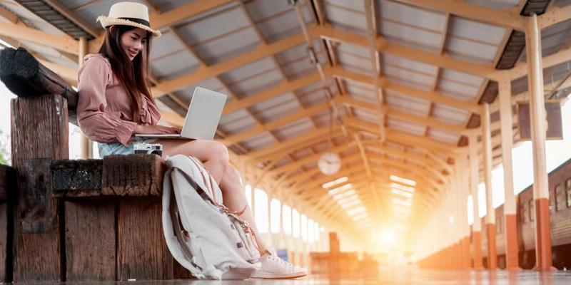 Frau mit Hut arbeitet am Laptop auf einem Bahnsteig