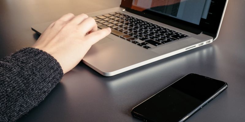 Hand auf der Tastatur eines Laptops, daneben ein Smartphone