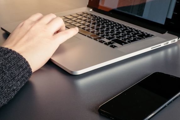 Hand auf der Tastatur eines Laptops, daneben ein Smartphone