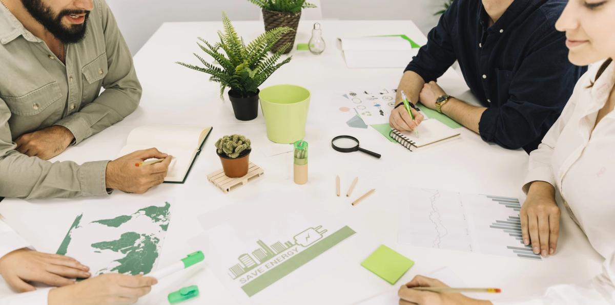 A group of people collaborating at a table with plants, notebooks, and charts, focusing on energy-saving ideas.