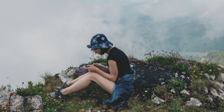 Frau mit Fischerhut sitzt entspannt auf einem Berggipfel im Nebel und schaut auf ihr Smartphone