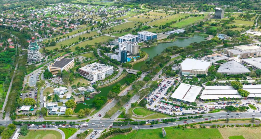 Aerial view of a mixed-use business district featuring office buildings, green spaces, roads, and a nearby residential area, surrounded by landscaped greenery and water features.