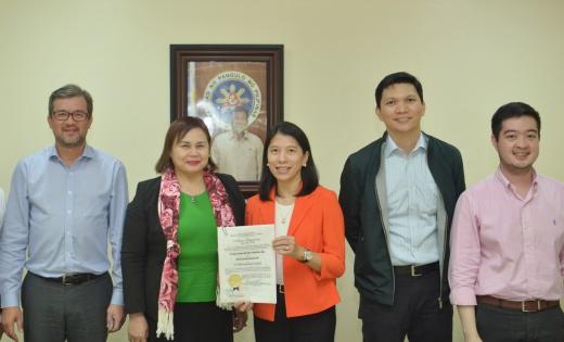 Six people stand together indoors, smiling for a group photo. The woman in the center holds up a certificate.
