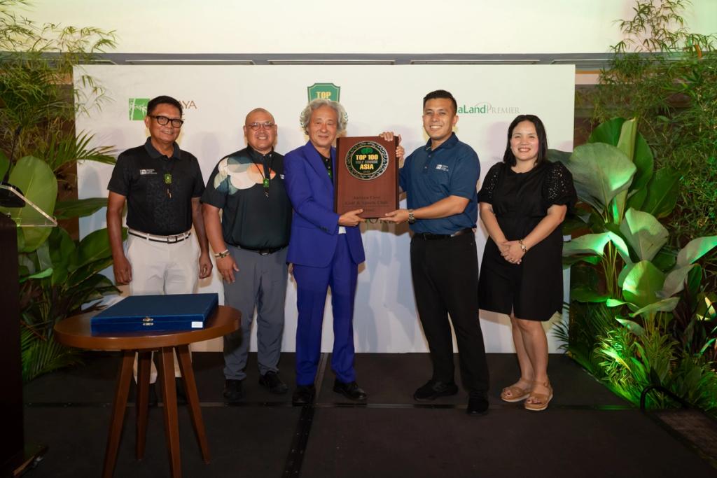 Five people stand together at an award ceremony. The central two individuals hold a large plaque featuring a green and gold seal. Everyone is smiling, and a branded backdrop with plants on either side suggests a formal recognition event related to sustainability or the environment.