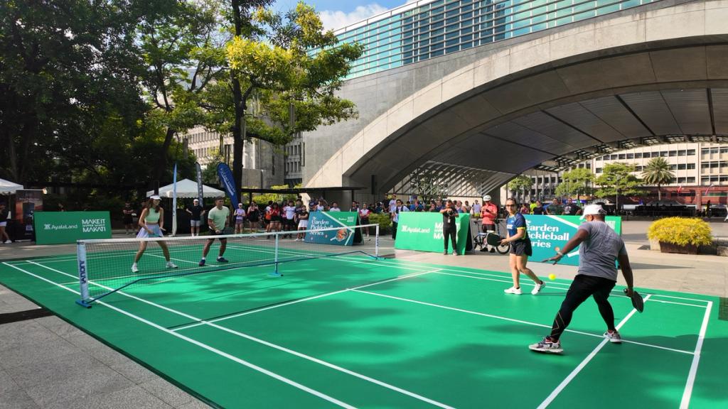 People playing pickleball on an outdoor court in Ayala Triangle Gardens in an urban setting with a modern bridge and city buildings in the background during a public sports event for Car Free Sundays at Ayala Avenue.