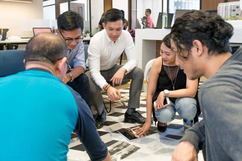 A diverse group of colleagues sitting together on the floor in a modern office, collaborating over printed materials and a tablet, engaged in a creative discussion.