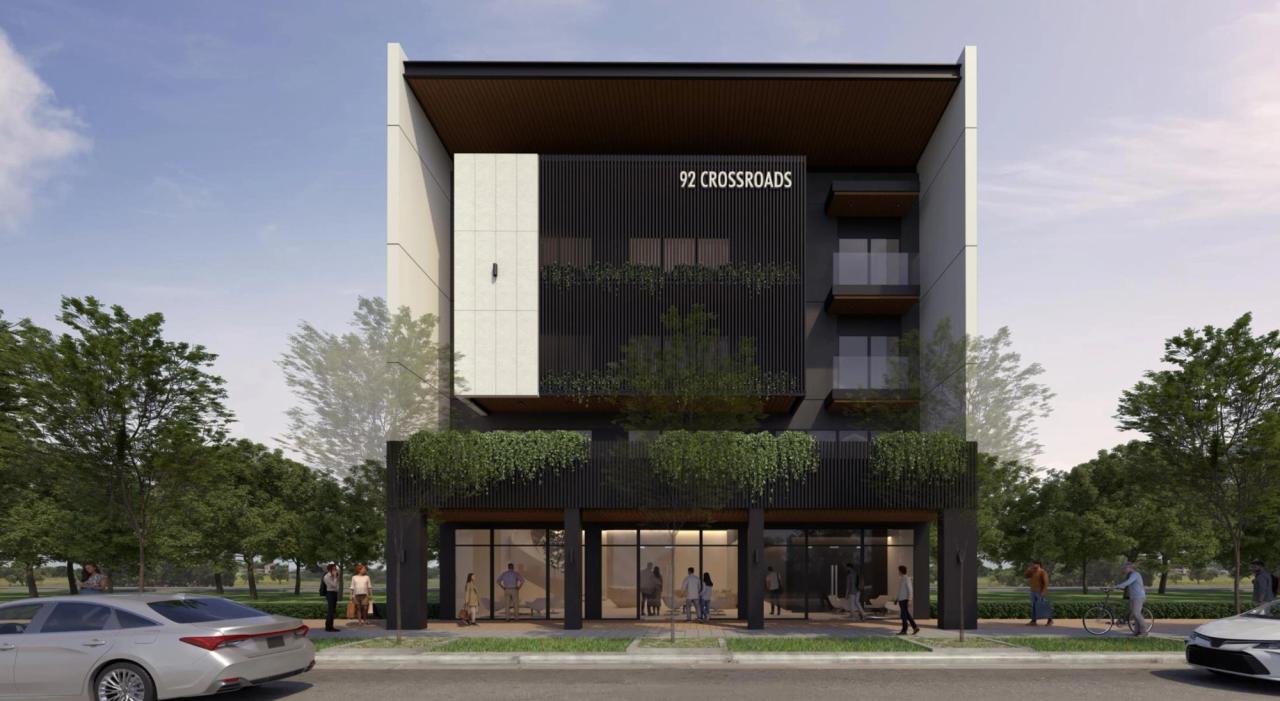 Front view of a modern mid-rise commercial building labeled “92 Crossroads,” featuring a minimalist façade with dark vertical slats, greenery accents on balconies, large glass storefronts on the ground floor, and people walking along a tree-lined street.