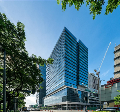 A modern glass office building stands on a busy street corner, surrounded by trees and other high-rise structures under a clear blue sky.