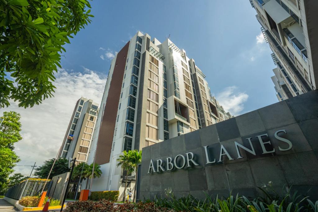 A modern mid-rise residential building with the name “Arbor Lanes” displayed on a stone sign at the entrance. The building features large windows and balconies, surrounded by landscaped greenery and palm trees under a bright blue sky.
