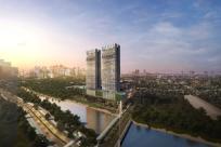 A scenic cityscape at sunset featuring a pair of modern high-rise residential towers beside a river, with a footbridge connecting to landscaped greenery and tree-lined pathways. The skyline of a bustling urban center rises in the background under a colorful sky.